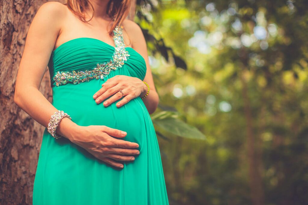 A pregnant woman wearing a green dress gently holds her belly in a serene outdoor setting.
