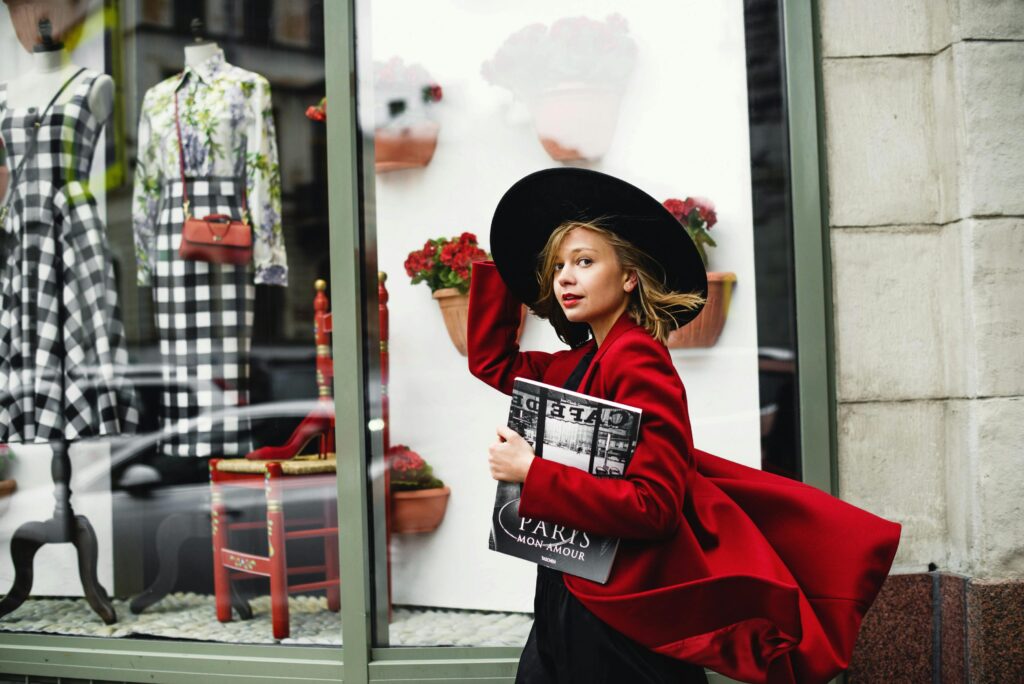 Fashionable woman in a red coat walks by a stylish boutique window, carrying a magazine.