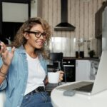 Smiling woman with glasses having a video call from home, enjoying a cup of coffee in her kitchen.
