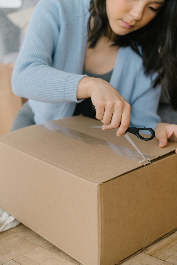 Asian woman using scissors to open a large cardboard box indoors.
